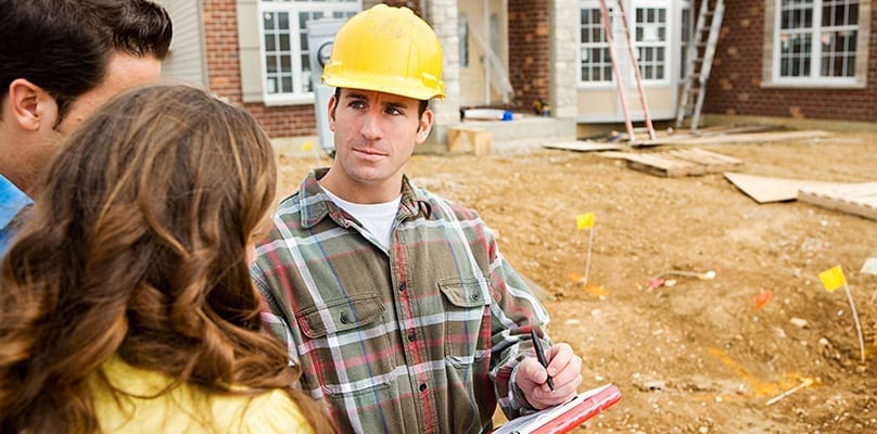 Contractor in a yellow hard hat with a clipboard talking with homeowners at a building site