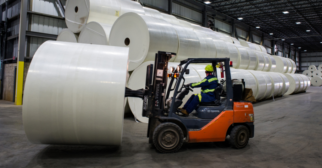 A forklift driver picking up a roll of fiberglass mat next to a long row of stacked fiberglass mats.