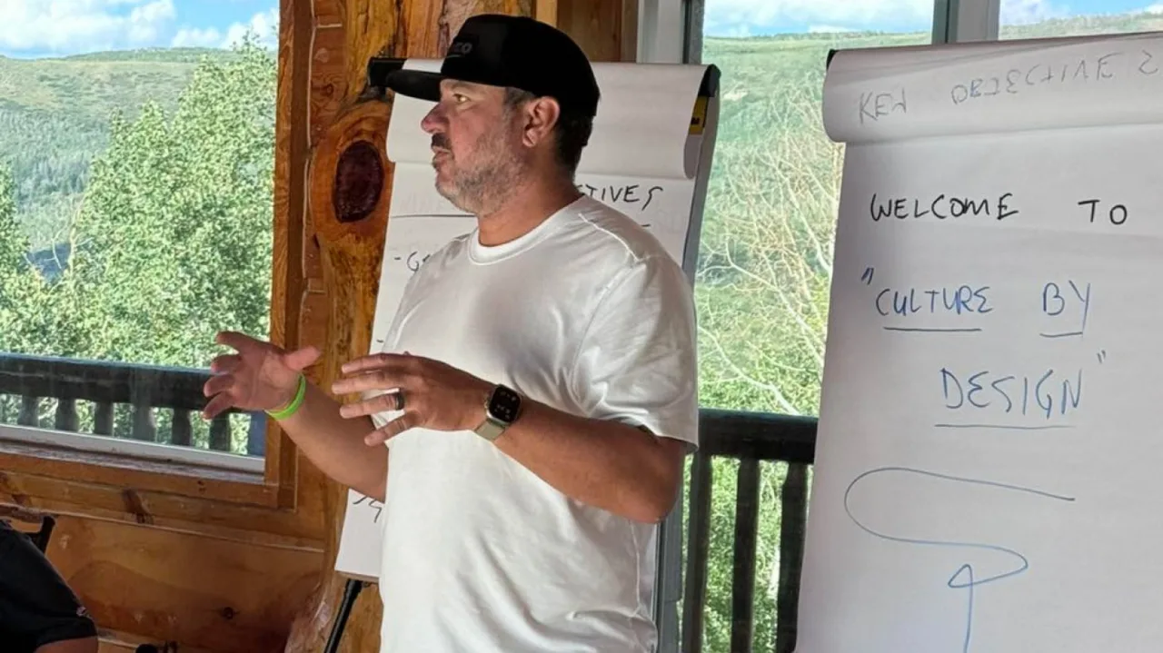 Un hombre con camiseta blanca habla con pasión sobre «Culture by Design» frente a rotafolios, con grandes ventanales que enmarcan árboles y colinas. Sus ideas reflejan los valores de ROOFPRO y cómo una cultura sólida beneficia al equipo y a los clientes.