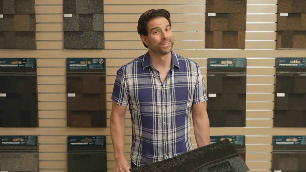 Man in a plaid shirt holding a roofing tile in a showroom, surrounded by samples on display walls.