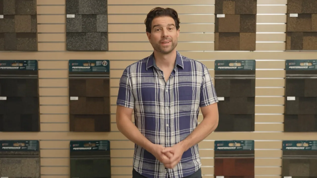 A man stands in a showroom wearing a plaid shirt, guiding homeowners through an array of IKO roofing samples on a beige slat wall.