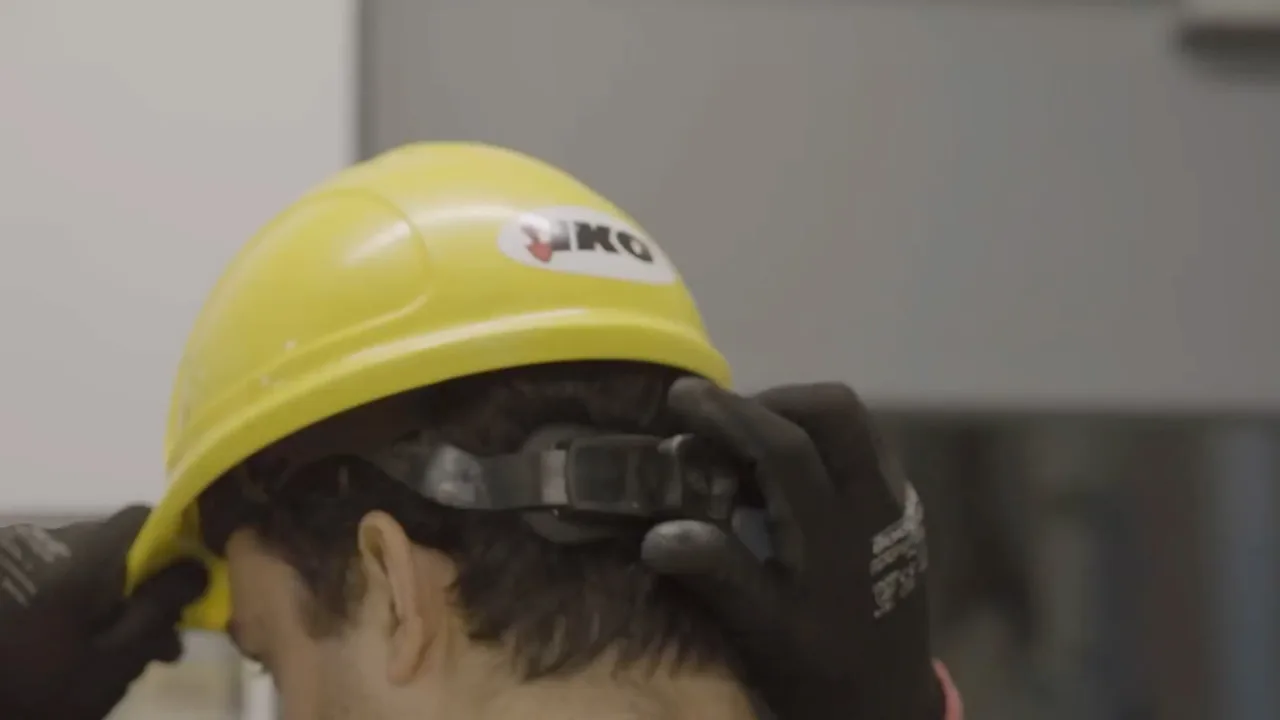 Person adjusting a yellow hard hat while wearing black gloves, with a blurred gray background.