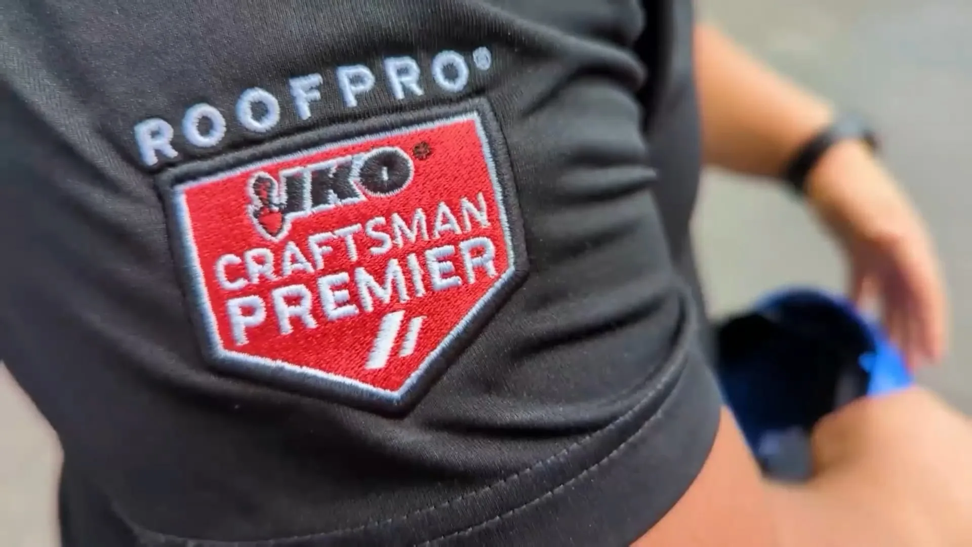Primer plano de una persona con camisa negra y un parche bordado, que destaca los beneficios de la insignia «ROOFPRO IKO CRAFTSMAN PREMIER» en llamativo texto rojo y blanco.