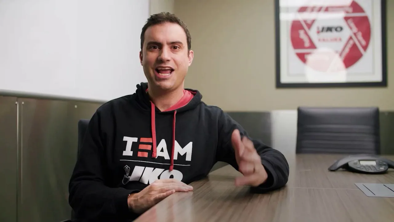 A person in a "TEAM" hoodie sits at the conference table, gesturing as they discuss the process. Wall art and a phone are visible in the background, adding to the professional ambiance.