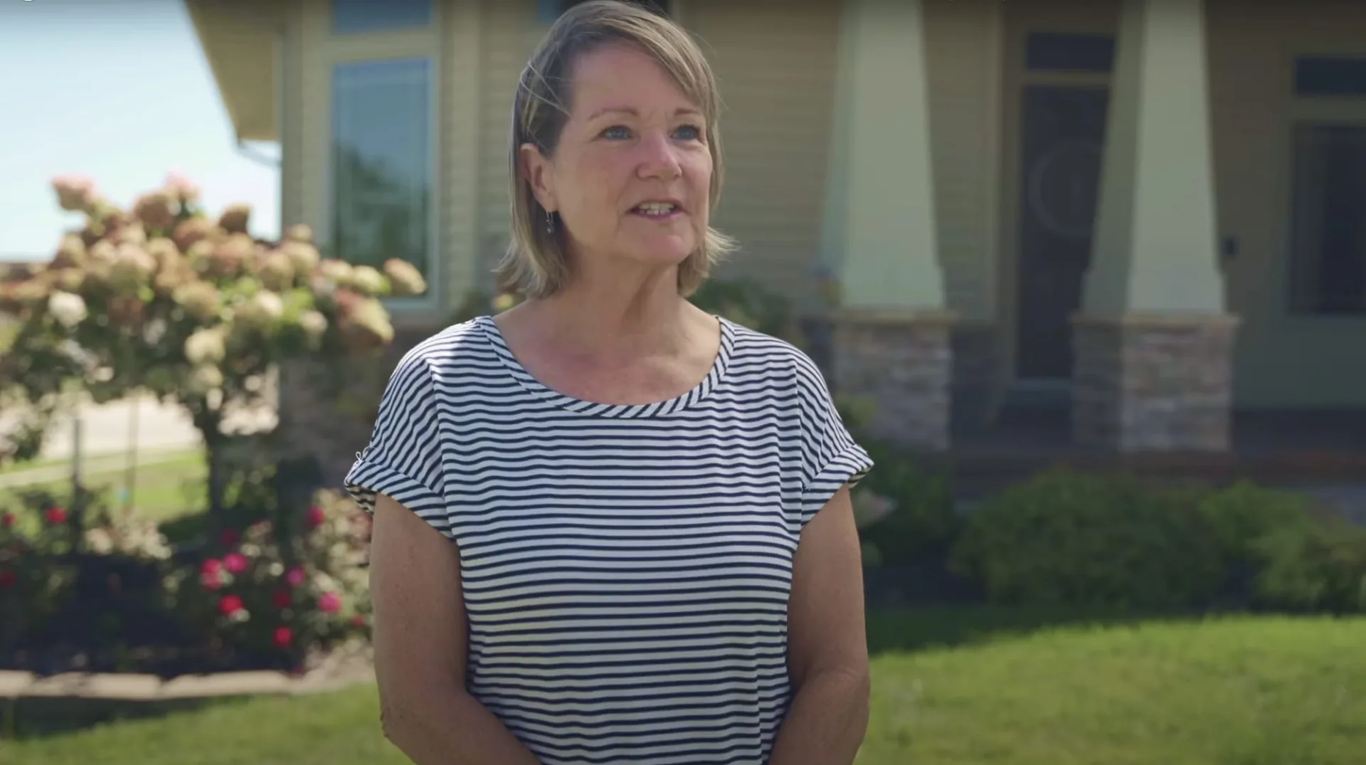Woman in a striped top speaking in front of a house with flowering shrubs
