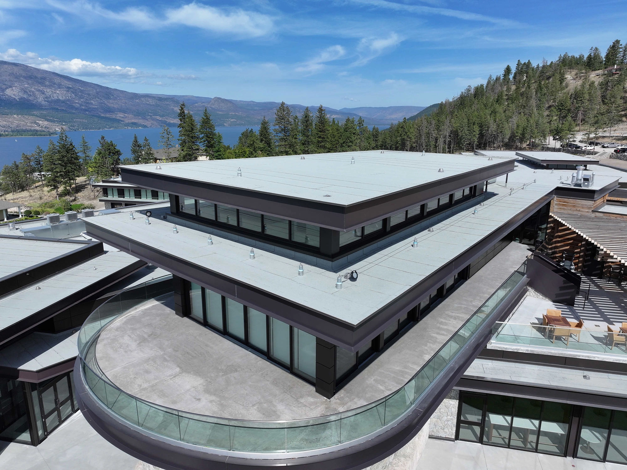 Vue aérienne d’un bâtiment moderne en bord de lac avec toit plat, balcon en verre courbe et montagnes en arrière-plan